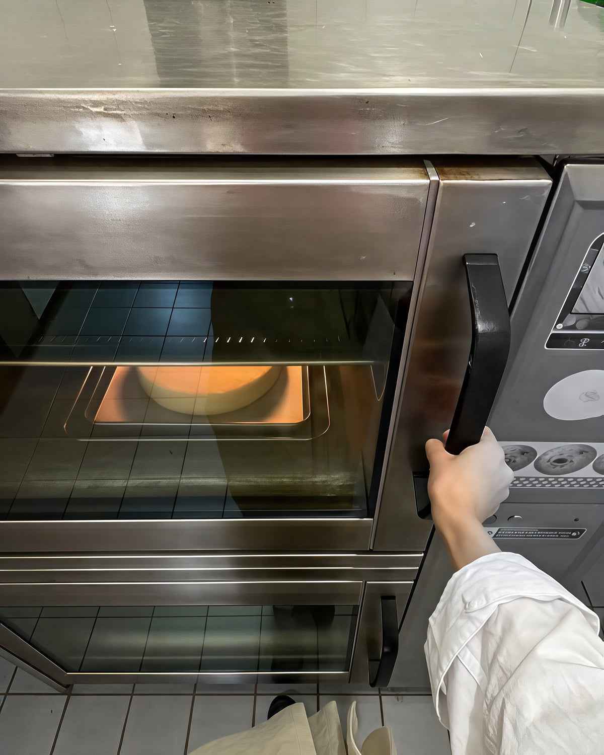 Over de schouder in een bakkerij: een hand opent de deur van de professionele oven, de cake staat binnen in het warme licht.